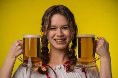 Neşeli genç bir kadın iki büyük bira bardağını canlı sarı arka planda tutarken geniş bir gülümseme sergiliyor..