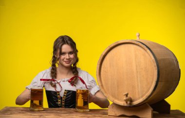 Geleneksel kıyafetli bir kadın, sarı arka planda tahta bir fıçının yanında iki bira bardağı tutarken gülümser..