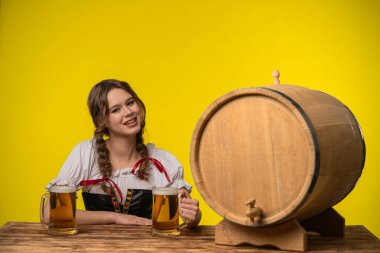 Geleneksel Bavyera kıyafeti giymiş bir kadın, elinde bira bardakları ve parlak sarı bir arka plana kurulmuş büyük bir tahta fıçıyla bir masada oturuyor..