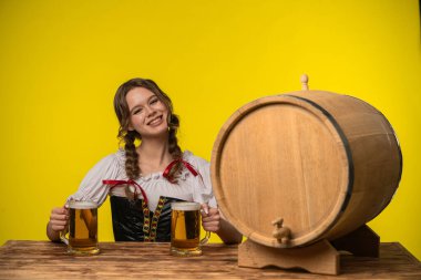 Geleneksel giyinmiş neşeli bir kadın, sarı arka planda, tahta bir fıçının yanında iki bira bardağı tutuyor..