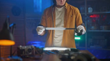 Eldiven ve turuncu ceket giyen bir kişi, klasik fotoğraf ekipmanları ve mavimsi ışıklandırma ile çevrili loş ışıklı bir odada bir film şeridini dikkatle tutar..