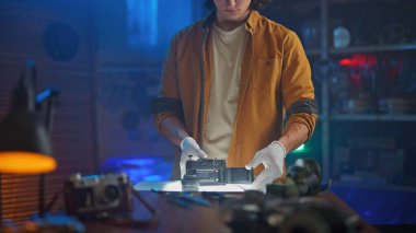 Bir kişi beyaz eldivenli eski bir kamerayı dikkatle inceliyor. Fotoğraf teçhizatı masada görünür..