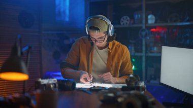 Kulaklıklı genç bir adam, yaratıcı bir şekilde aydınlatılmış stüdyo ortamındaki bir masada yoğun bir şekilde işine konsantre oluyor..