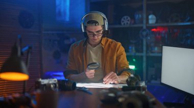 Kulaklık ve şapka takan genç bir adam loş bir odada büyüteçle belgeleri dikkatle inceliyor..