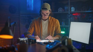 Günlük kıyafetli bir kişi, düşük ışık ortamında kamera ve ekipmanlarla film negatiflerini analiz ediyor..