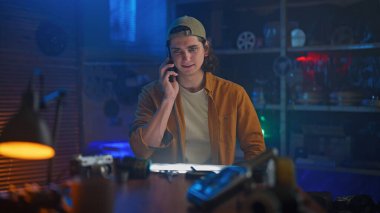 Günlük giysiler içinde genç bir adam, bir atölyede loş ışıklandırma ile telefonuyla konuşuyor, yaratıcılık hissi uyandırıyor..