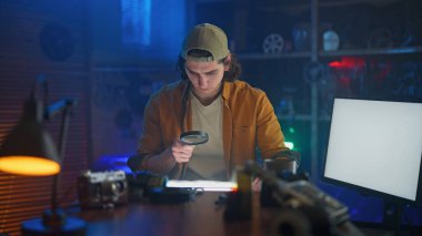 Rahat bir odada genç bir adam çeşitli aletlerle çevrili bir büyüteçle fotoğrafları inceliyor..