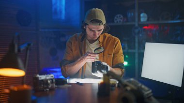 Genç bir adam, sıcak ve yaratıcı bir stüdyo ortamında elinde büyüteçle kamerayı incelemeye odaklanıyor..