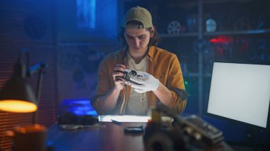 Genç bir adam, teknik ekipman ve çevre aydınlatması ile çevrili bir atölyedeki eski bir kamerayı dikkatle inceliyor..