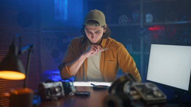 Odaklanmış genç bir adam çalışmalarını loş bir stüdyoda yaratıcı aletlerle dolu bir masada düşünür..