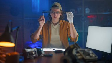 Şapkalı ve eldivenli genç bir adam stüdyo ortamında bir kamera filmini inceliyor, analog fotoğrafçılığa ilgi gösteriyor..
