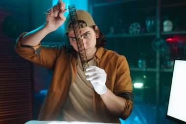 Odaklanmış genç bir dedektif loş bir odada eldivenli ve kepli bir film şeridini inceliyor..