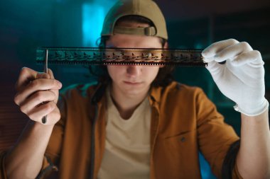 Eldiven takan bir adam eski bir film şeridini inceliyor, profesyonel bir ortamda dikkatli kullanılması gerektiğini vurguluyor..