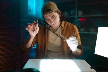 Genç bir insan loş bir odada parlak ışık altında bir film şeridini dikkatle inceler, şapka ve eldiven giyer..