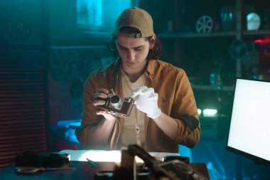 Eldiven giyen genç bir adam atölyedeki eski bir kamerayı inceliyor. Hassasiyet ve özeni vurguluyor..