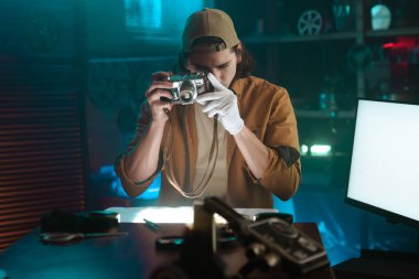 Stüdyo ortamında eski bir kameraya odaklanmış sıradan bir kıyafet ve eldiven giymiş bir adam. Muhtemelen fotoğraf çekimi ya da adli tıp incelemesi için..