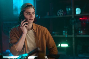 Sıradan kıyafetler içindeki genç bir adam loş ışıklı bir atölyenin içinde telefonla konuşuyor ve sıradan bir ortam sergiliyor..