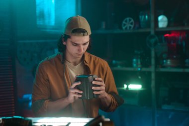 Günlük kıyafetli genç bir adam loş bir atölye ortamındaki ekipmanları dikkatle inceliyor..