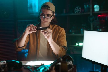 Şapkalı ve gözlüklü genç bir adam loş ışıklı karanlık bir odada film negatifini dikkatle inceliyor. Bu da fotoğrafçılığa ya da film yapımına odaklanılmasını öneriyor..