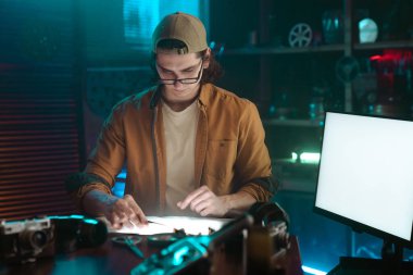 Gözlük ve şapka takan genç bir adam, çağdaş bir çalışma alanında tasarım projeleri üzerinde çalışıyor..