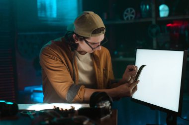 Bir adam karamsar, loş bir odada negatif bir filmi yakından inceler. Ortam yaratıcı, odaklanmış bir çalışma alanı..