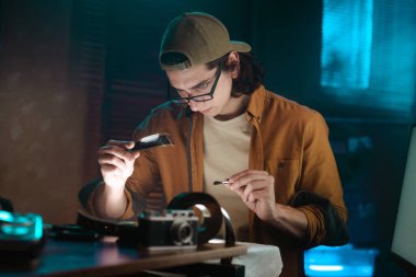 Genç bir adam loş aydınlatılmış bir odada dikkatle bir film şeridini inceliyor. Detaylara odaklanma ve dikkat gösteriyor..