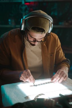 Odaklanmış bir teknisyen, loş bir ortamda gözlük ve kulaklık takarak ışık masasındaki film şeritlerini inceliyor..