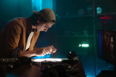 Stüdyo ortamında genç bir adam, bir objeyi dikkatle incelemeye odaklanmış. Düşük ışık ve yaratıcı ortam.