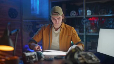 Genç bir adam, teknoloji ve aletlerle çevrili loş ışıklı yaratıcı bir stüdyoda film şeridini inceliyor..