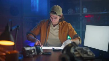 Genç bir adam, teknoloji ve aletlerle çevrili loş ışıklı yaratıcı bir stüdyoda film şeridini inceliyor..