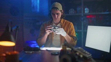 Genç bir adam, teknik ekipman ve çevre aydınlatması ile çevrili bir atölyedeki eski bir kamerayı dikkatle inceliyor..