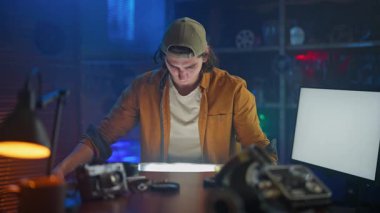 Genç bir adam, teknoloji ve aletlerle çevrili loş ışıklı yaratıcı bir stüdyoda film şeridini inceliyor..