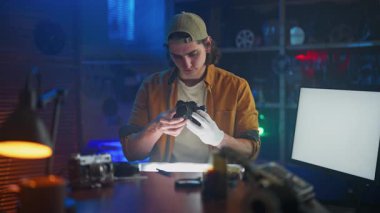 Genç bir adam, teknik ekipman ve çevre aydınlatması ile çevrili bir atölyedeki eski bir kamerayı dikkatle inceliyor..