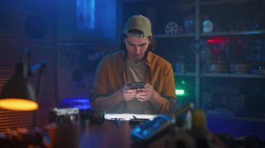 Günlük giysiler içinde genç bir adam, bir atölyede loş ışıklandırma ile telefonuyla konuşuyor, yaratıcılık hissi uyandırıyor..