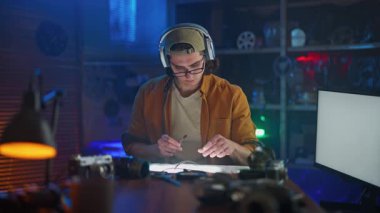 Kulaklıklı genç bir adam, yaratıcı bir şekilde aydınlatılmış stüdyo ortamındaki bir masada yoğun bir şekilde işine konsantre oluyor..