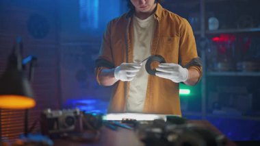 Eldiven ve turuncu ceket giyen bir kişi, klasik fotoğraf ekipmanları ve mavimsi ışıklandırma ile çevrili loş ışıklı bir odada bir film şeridini dikkatle tutar..