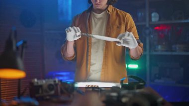 Eldiven ve turuncu ceket giyen bir kişi, klasik fotoğraf ekipmanları ve mavimsi ışıklandırma ile çevrili loş ışıklı bir odada bir film şeridini dikkatle tutar..