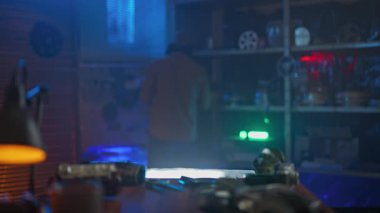 Eldiven ve turuncu ceket giyen bir kişi, klasik fotoğraf ekipmanları ve mavimsi ışıklandırma ile çevrili loş ışıklı bir odada bir film şeridini dikkatle tutar..