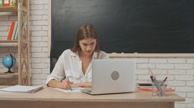 Okul öğretmeni sınıfta öğrencilere konuşuyor. Sınıfın masasındaki yazı tahtasının önünde oturan genç bayan üniversite öğretmeni öğrencilere laptoptaki video çağrısından ders veriyor. Eğitim kavramı.