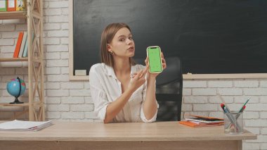 Okul öğretmeni sınıfta çalışır. Sınıftaki masada oturan genç bayan üniversite öğretmeni akıllı telefon kromu yeşil ekran tutuyor ve baş parmağını gösteriyor..