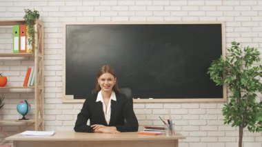 Okul öğretmeni sınıfta çalışır. Sınıfta masanın önünde oturan genç bayan üniversite öğretmeni kameraya bakıp gülümsüyor, olumlu ifade. Eğitim kavramı.