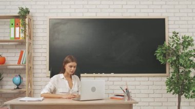 Okul öğretmeni sınıfta öğrencilere konuşuyor. Sınıfın masasındaki yazı tahtasının önünde oturan genç bayan üniversite öğretmeni öğrencilere bilgisayar kullanarak video araması yaparak ders veriyor. Eğitim kavramı.