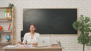 Okul öğretmeni sınıfta öğrencilere konuşuyor. Sınıfın önündeki masada oturan genç bayan üniversite öğretmeni, ilkokul çocuklarına bilgisayar kullanarak ders veriyor. Eğitim kavramı.