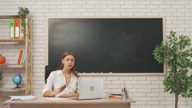 Okul öğretmeni sınıfta öğrencilere konuşuyor. Sınıfta masa başında yazı tahtası önünde oturan kadın üniversite öğretmeni çocuklara laptoptaki video çağrısından ders veriyor. Eğitim kavramı.
