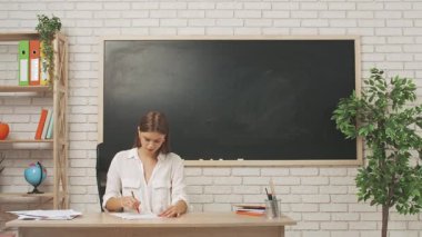 Okul öğretmeni sınıfta çalışır. Sınıftaki genç bayan üniversite öğretmeni kırmızı kalemli sınav kağıtlarını karatahtanın önünde okuyor. Eğitim kavramı.