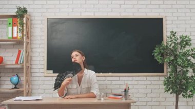 Okul öğretmeni sınıfta çalışır. Sınıftaki genç bayan kolej öğretmeni yazı tahtasının önünde el sallıyor içme suyu içiyor, sıcaklıkla başa çıkıyor, yorgun bir ifade takınıyor. Eğitim kavramı.