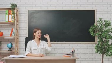 Okul öğretmeni sınıfta çalışır. Sınıftaki masada oturan genç bayan üniversite öğretmeni boş alanı işaret eden yazı tahtasının önünde başparmağını kaldırıyor. Eğitim kavramı.