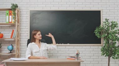 Okul öğretmeni sınıfta çalışır. Sınıftaki masada oturan genç bayan üniversite öğretmeni yazı tahtasının önünde boş yerleri işaret ederek kameraya gülümsüyor. Eğitim kavramı.