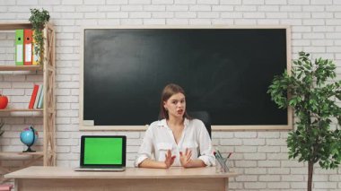 Okul öğretmeni sınıfta öğrencilere konuşuyor. Sınıfın önündeki masada oturan genç bayan kolej öğretmeni öğrencilere dersi açıklıyor, dizüstü bilgisayar krom anahtar yeşil ekran. Eğitim kavramı.