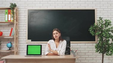 Okul öğretmeni sınıfta öğrencilere konuşuyor. Sınıfın masasındaki yazı tahtasının önünde oturan kadın öğretmen, öğrencilere dersi açıklıyor, dizüstü bilgisayar krom anahtar yeşil ekranı gösteriyor. Eğitim kavramı.